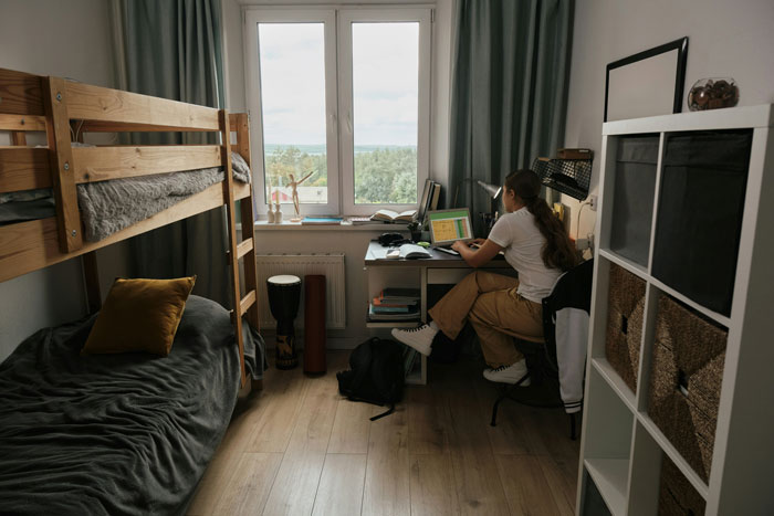 Teen in cozy bedroom with bunk bed and desk, using laptop, faced with parents' request to move to basement for relatives.