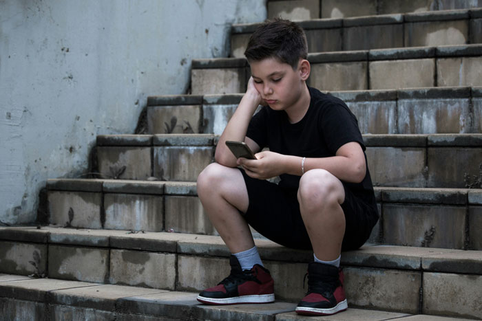 Teen in a dingy basement, sitting on stairs, looking at phone, wearing casual clothes and red sneakers.