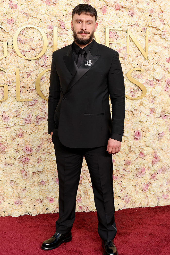 Man in black suit on red carpet at Golden Globes 2025, posing against a floral backdrop.