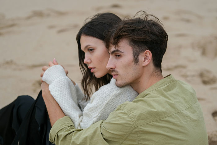 Couple sitting on a sandy beach, embracing closely, capturing a romantic engagement moment.