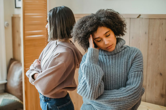 Two women having a disagreement, one looking upset, in a room with wooden paneling.