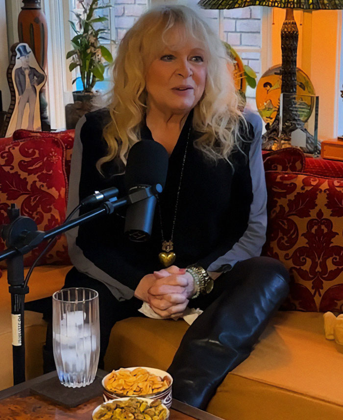 Blonde woman speaking into a microphone on a red sofa, with snacks and a drink nearby.