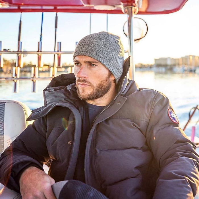 Clint Eastwood's son in a winter jacket and beanie, sitting on a boat, looking contemplative during a sunny day.