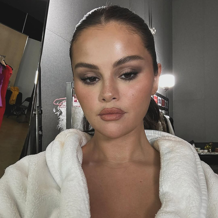 Selena Gomez in a white robe with smoky eye makeup, looking reflective under bright lights.