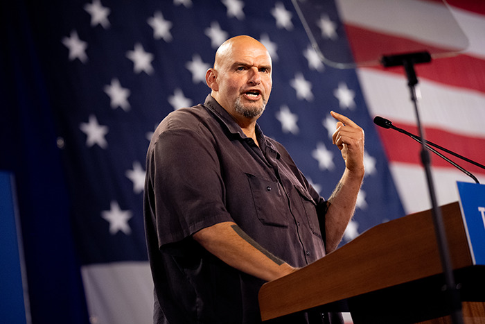 Senator speaking at a podium during a presidential inauguration, wearing casual clothing in front of an American flag.