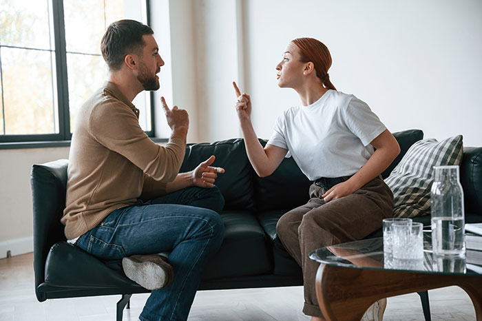 Man and woman sitting on a sofa, engaged in a heated discussion, raising fingers as they argue about rent.