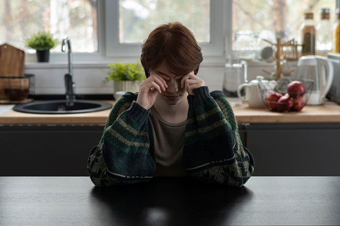 Woman sitting at a kitchen table, wearing a green sweater, looking down, related to spoiled brat controversy.