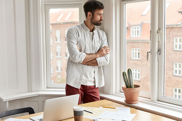 Man in a bright room, arms crossed, gazing out the window, with a desk, laptop, and cactus in the foreground.