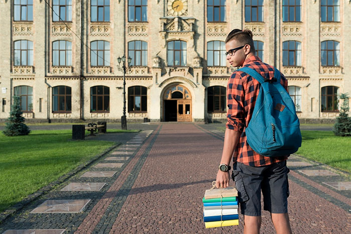 Young man with a backpack holding books, standing in front of a historical building on a sunny day.