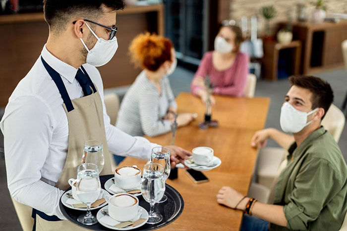 Waiter serving drinks to three people at a cafe, all wearing face masks.