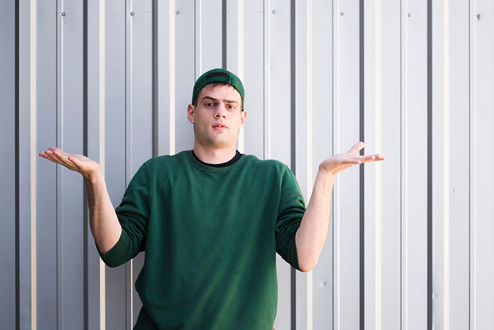 Man in green sweatshirt and cap shrugging in confusion, standing against a metal wall.