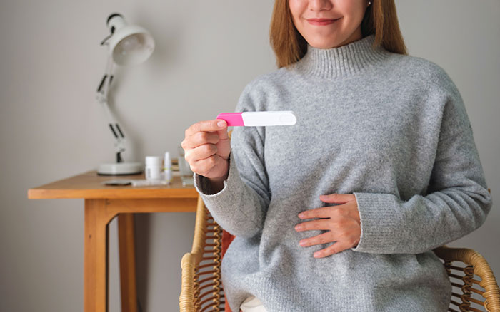 Woman holding a pregnancy test with a smile, touching her belly, announcing pregnancy.