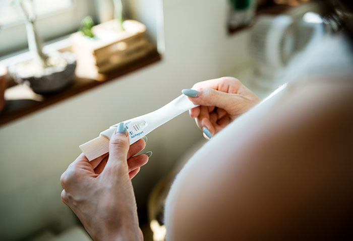 Woman holding a positive pregnancy test by a window, capturing a moment of anticipation and emotion.