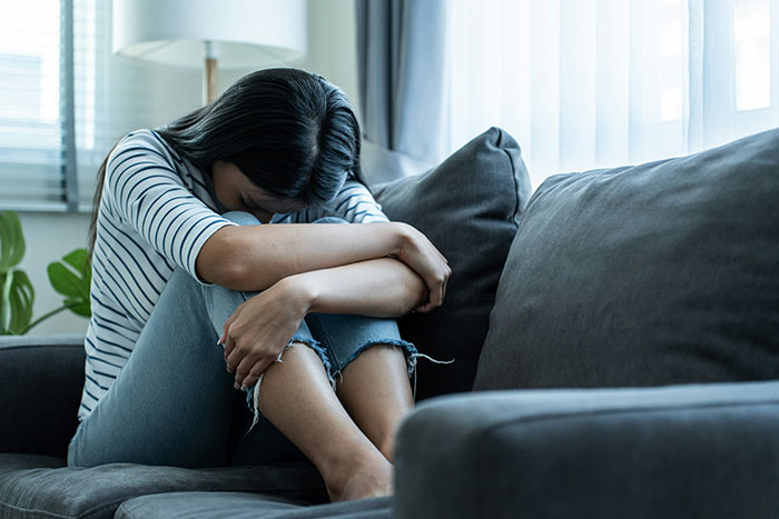 Woman in distress sitting on a couch, concealing her face with knees, struggling with relationship issues.