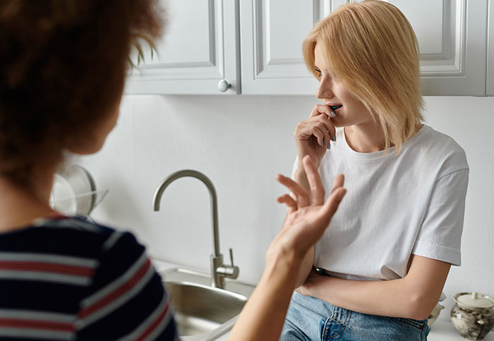 Two women in a kitchen have a discussion about a baby, one gesturing and the other looking thoughtful.