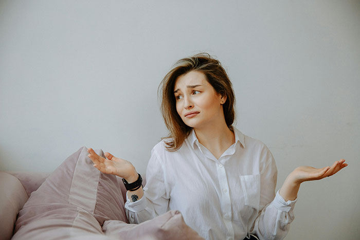 A woman in a white shirt sitting on a couch, shrugging, symbolizing a family conflict over an heirloom ring.