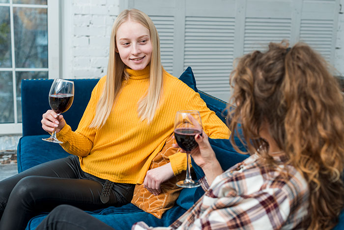 Two sisters having a conversation on a blue couch, each holding a glass of red wine, discussing an heirloom ring.