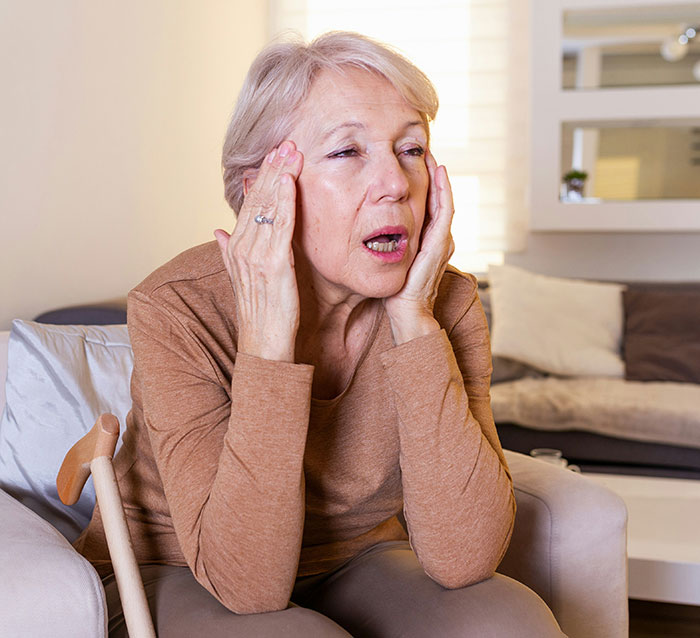 Elderly woman sitting on a sofa, appearing surprised, with a cane beside her.