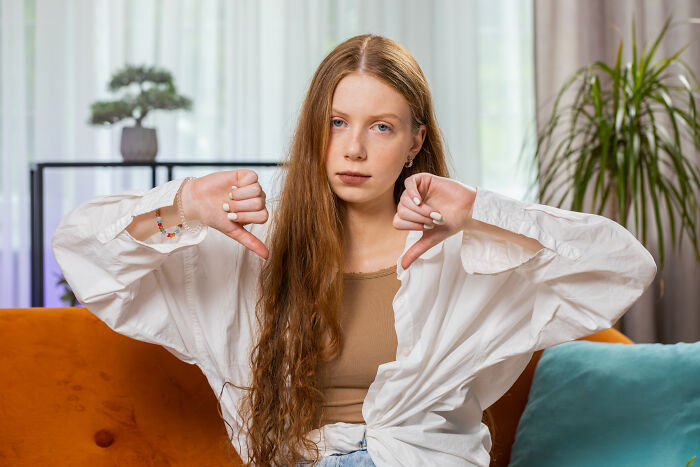 A young girl sitting on an orange couch with thumbs down, representing disapproval in a family home setting.