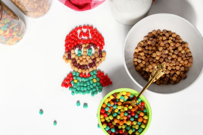 Colorful cereal art depicting a cartoon character using fresh ingredients on a white surface.