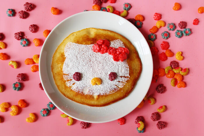 Playful food art of a pancake decorated like a cartoon character surrounded by colorful cereal on a pink background.