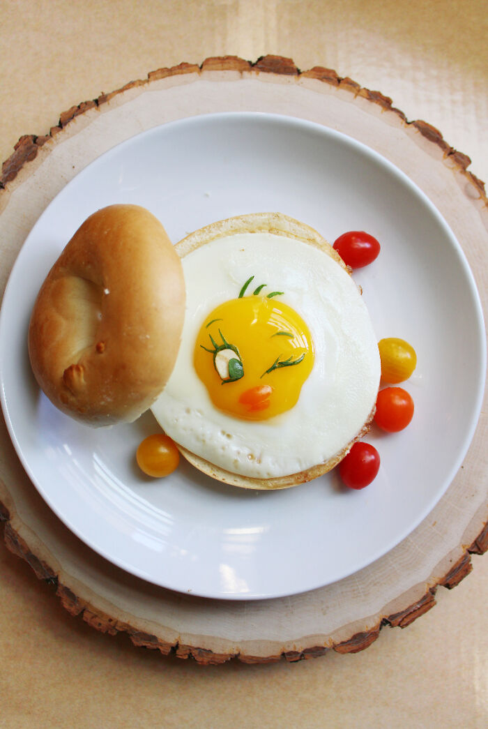 Playful food art inspired by cartoons, featuring a fried egg as a character on a plate with a bagel and cherry tomatoes.