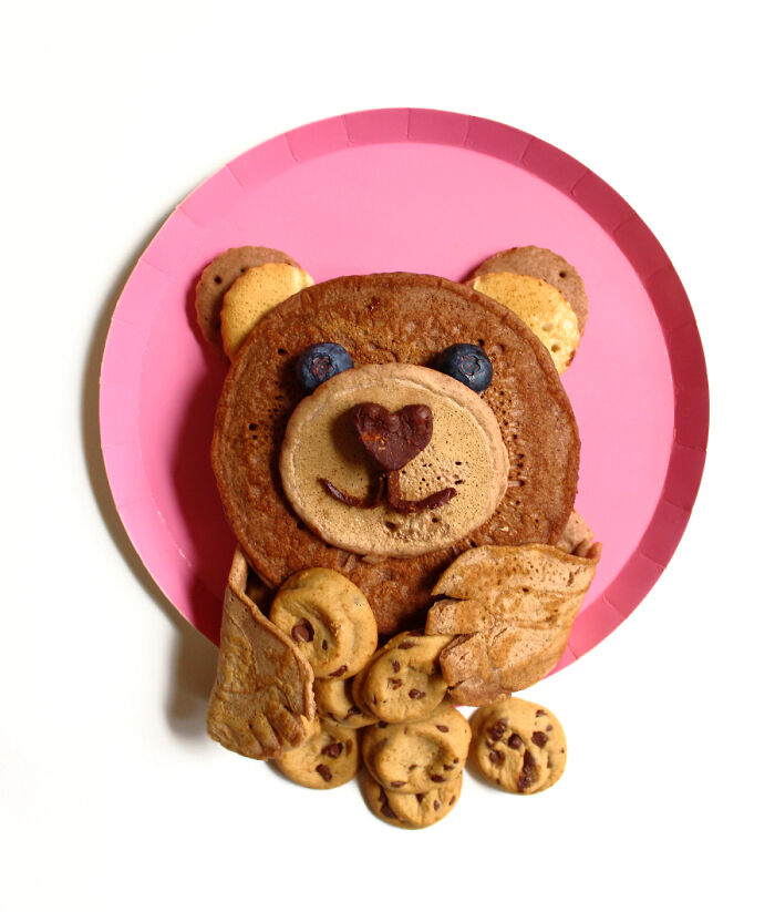 Playful food art of a bear made with fresh ingredients, including pancakes and cookies, on a pink plate.