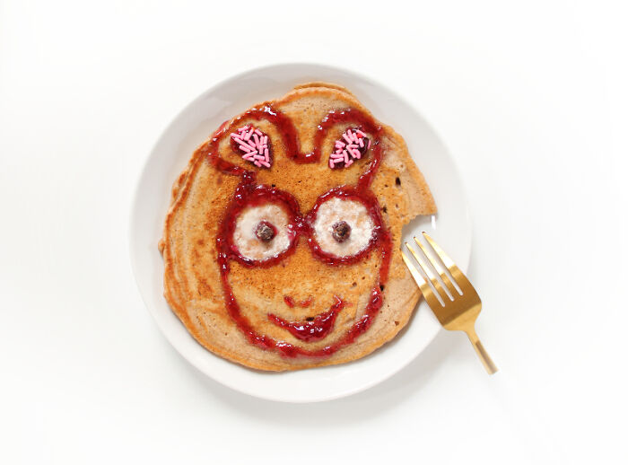 Pancake decorated with playful food art resembling a cartoon face, made using fresh ingredients, and garnished with pink sprinkles.