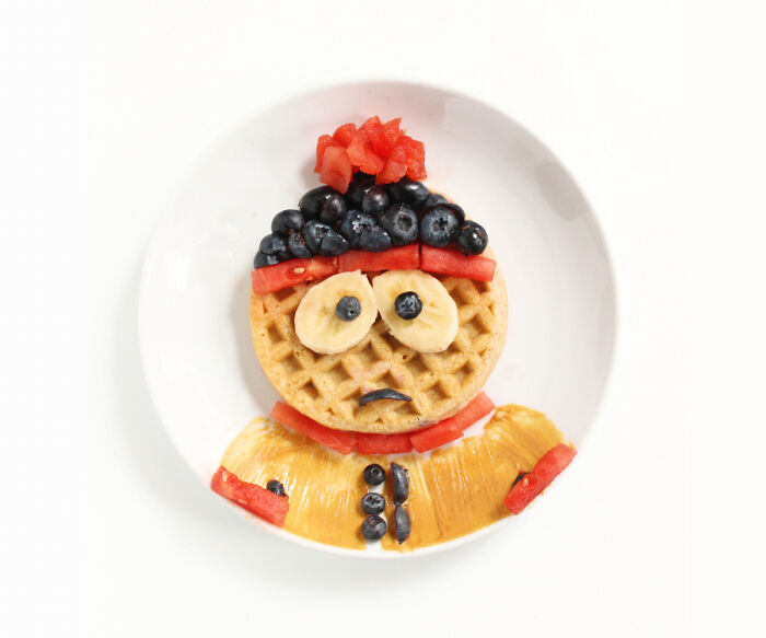 Cartoon-inspired playful food art made with fresh fruits on a waffle, resembling a character on a white plate.