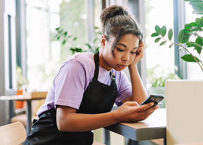 A staff member in a cafe, wearing a purple shirt and apron, looks at a phone.