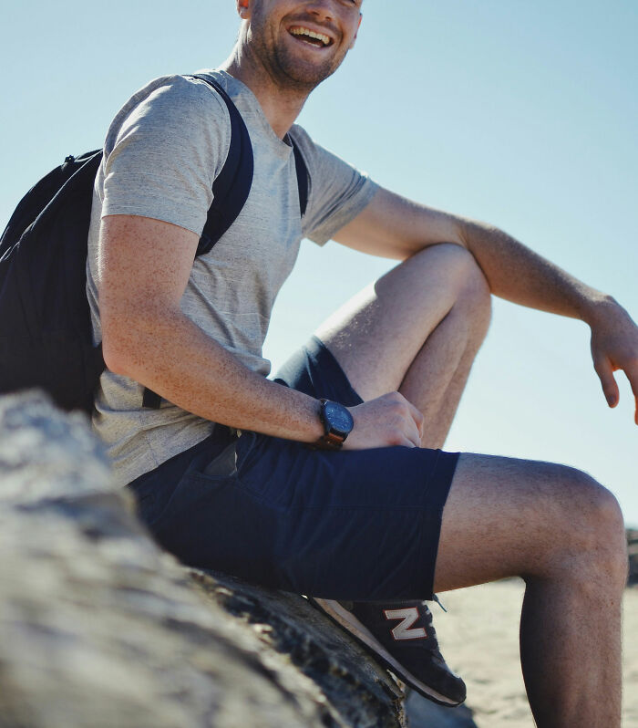 Man sitting on a rock, smiling while wearing casual clothes and a backpack, outdoors under clear skies.