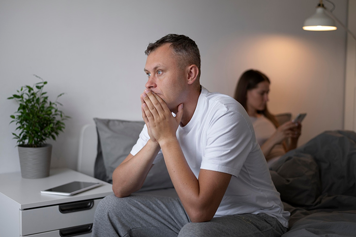 Man sitting thoughtfully on bed, woman using phone in background; concept of friends backing out before success.