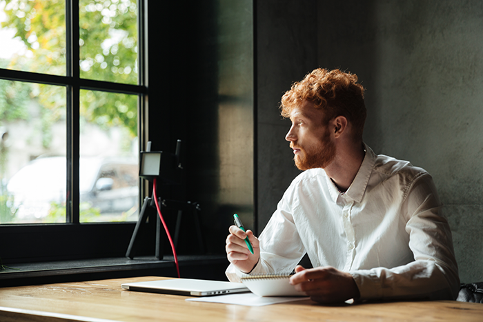 Man in a white shirt, sitting at a desk with notebook, looking thoughtfully out a window.