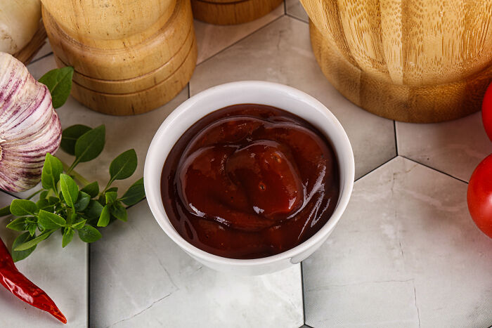 Bowl of ketchup on a table with garlic, herbs, and tomatoes, illustrating a playful revenge theme.