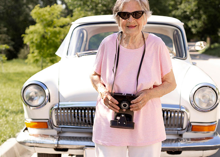 Elderly woman smiling with a camera, standing in front of a classic car on a sunny day.