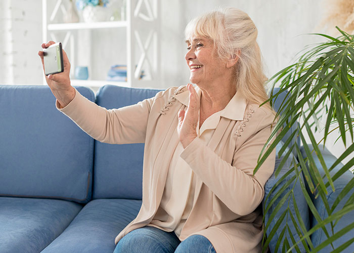 Older person taking a selfie on a smartphone, smiling and waving while sitting on a blue sofa.