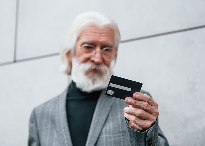 Elderly man with a beard in a gray suit looking at a credit card, highlighting teaching grandparents skills.