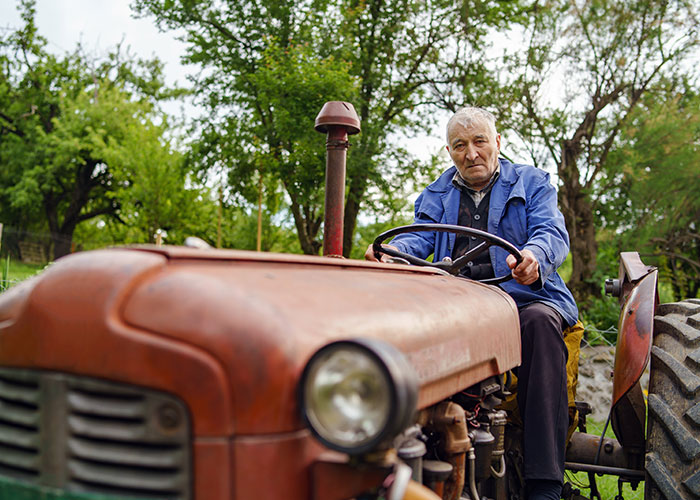 Older person in blue jacket seated on a vintage tractor, surrounded by trees, illustrating intergenerational learning.