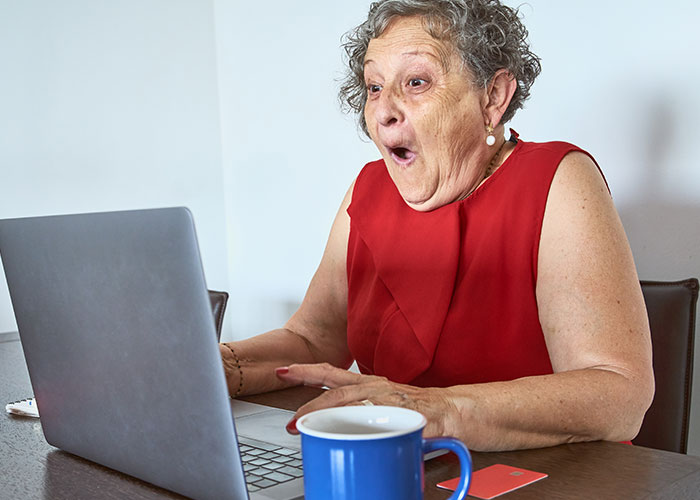 Older person in a red shirt joyfully reacts to a laptop while sitting at a table with a blue mug.