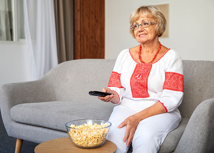 Older woman on a gray sofa with a remote and popcorn, illustrating teaching moments with grandparents.