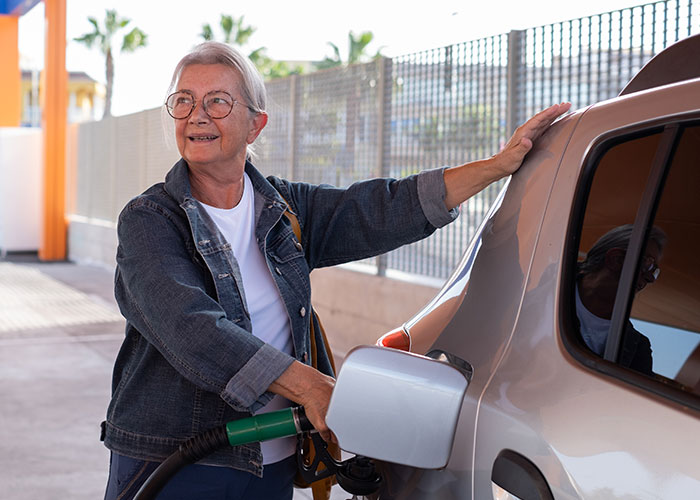 Older person refueling car, smiling, wearing glasses and denim jacket, teaching moment at gas station.