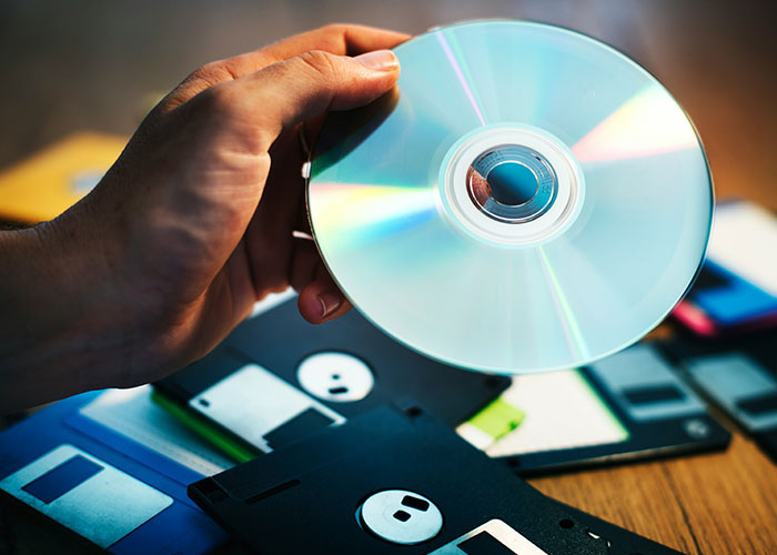 Hand holding a CD above floppy disks, showcasing older technology and lessons shared between generations.