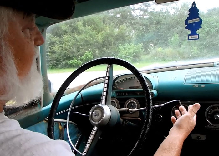 Older person teaching grandparent in a car, gesturing at a retro dashboard with trees visible through the windshield.