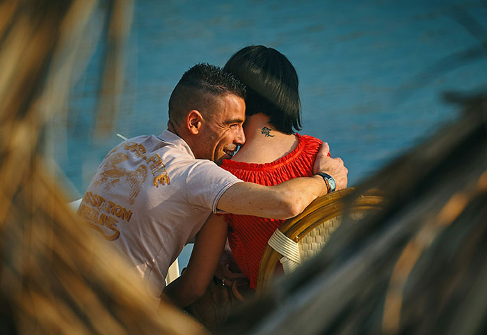 Man sitting next to a woman by the water, with arm around her shoulders, capturing a cozy moment.