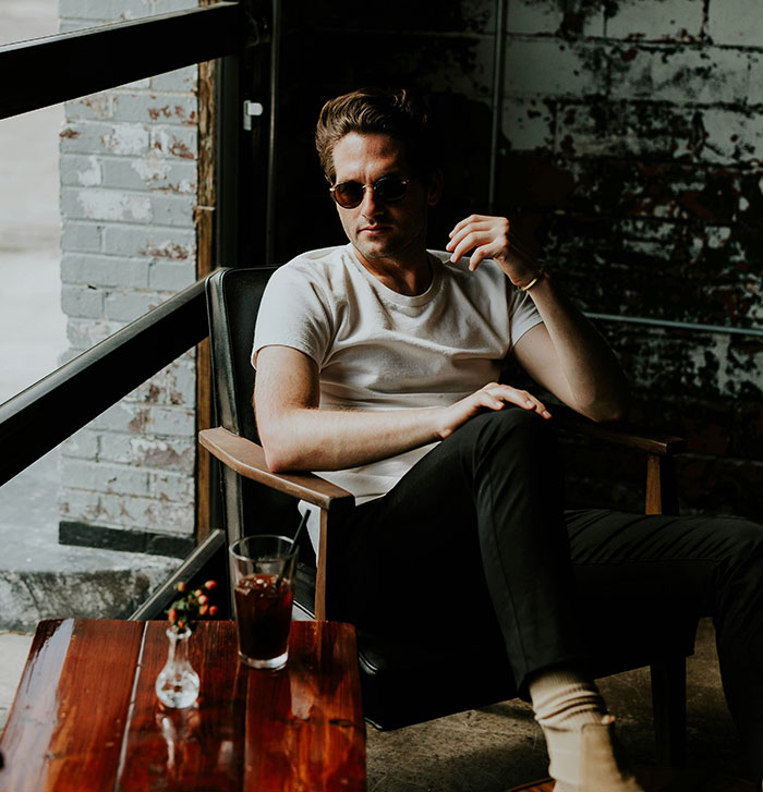 Man in a cozy cafe wearing sunglasses, sitting next to a rustic wooden table, exuding a laid-back vibe.