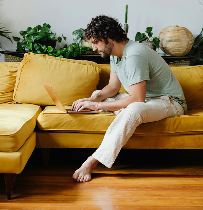 Man using a laptop on a mustard sofa, surrounded by plants in a casual setting.