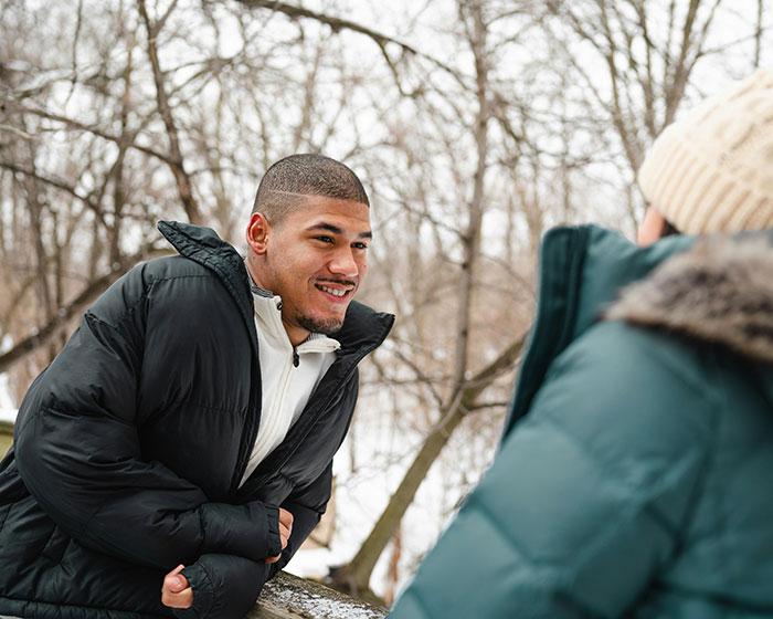 Man in a black jacket leaning on a snowy railing, engaging in conversation, embodying social interactions outdoors.