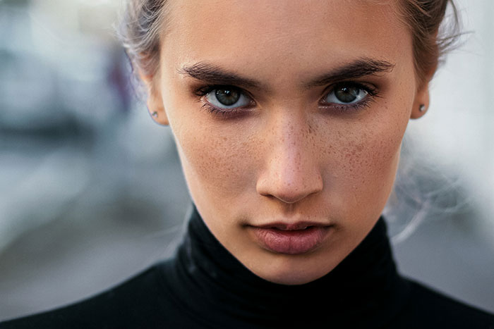 A woman with a serious expression, freckles visible, wearing a black top in an outdoor setting.