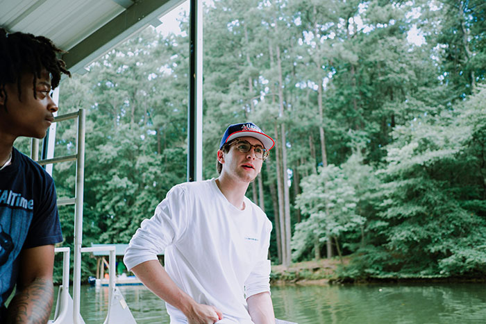 Two men sitting on a boat beside each other, surrounded by lush trees, engaging in casual conversation.