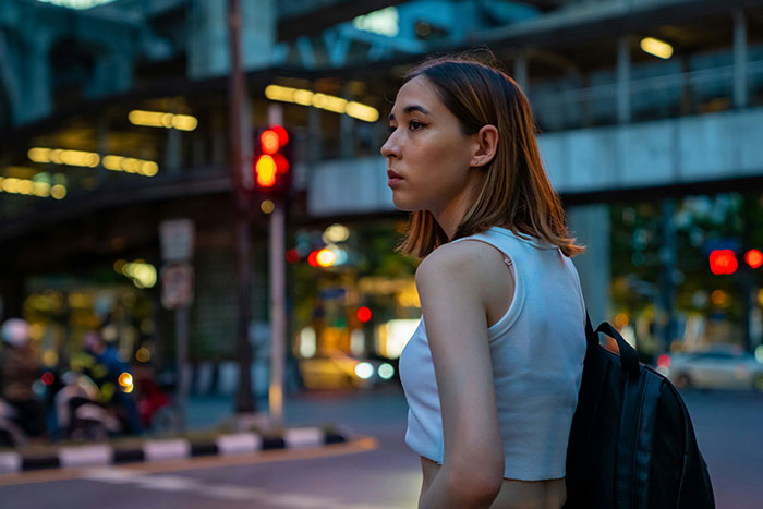 Woman standing at a city intersection at dusk, looking contemplative with a backpack, under streetlights.